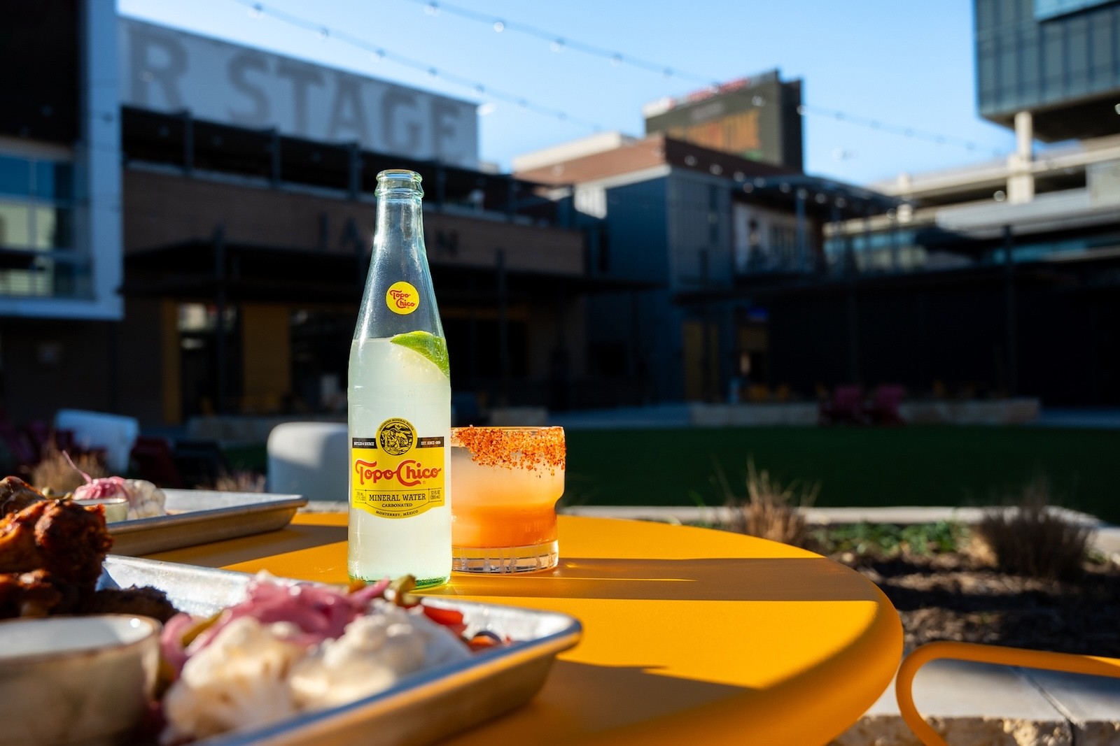 Food and drinks arranged on an outdoor table at Toyota Music Factory, with a Topo Chico bottle and a stage in the background.