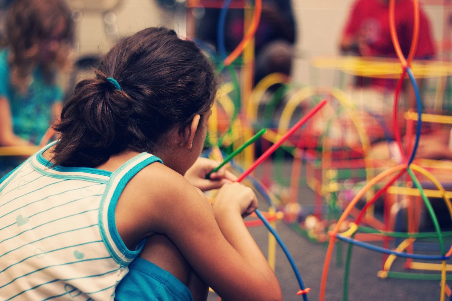 A imagem mostra uma criança envolvida em uma atividade lúdica com brinquedos educativos coloridos. Ela parece concentrada, manipulando bastões ou fios, cercada por um ambiente repleto de formas e cores vibrantes. Esse tipo de atividade geralmente é usado para estimular a criatividade, coordenação motora e habilidades de resolução de problemas nas crianças, proporcionando um ambiente de aprendizado interativo. A atmosfera parece de um espaço de brincadeiras ou uma sala de atividades em uma escola ou centro educativo.