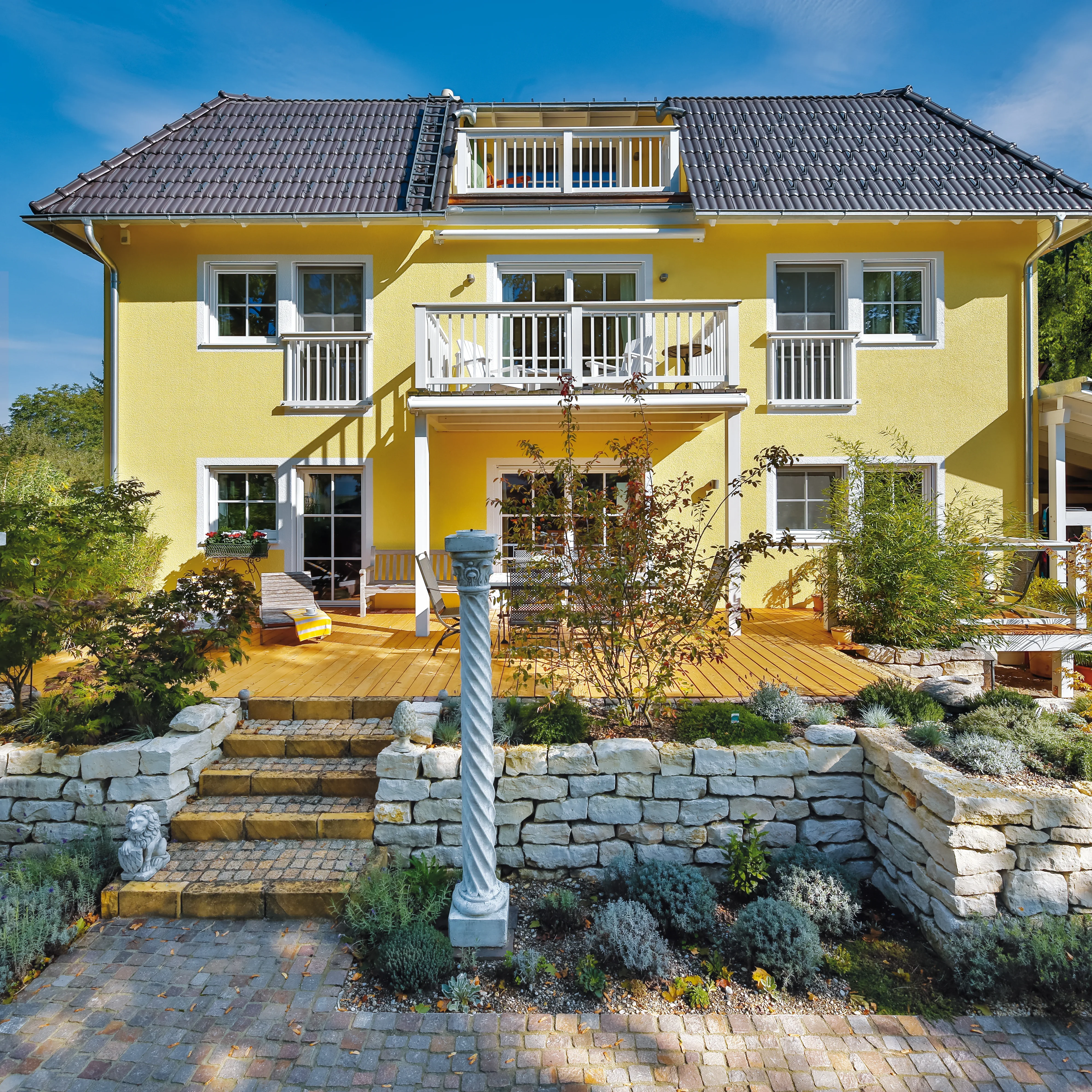 Gelbes Einfamilienhaus mit großzügiger Holzterrasse und Gartenanlage mit Steineinfassungen und Zierpflanzen.