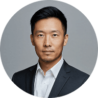 An Asian man wearing a formal suit and tie, standing confidently with a neutral expression.