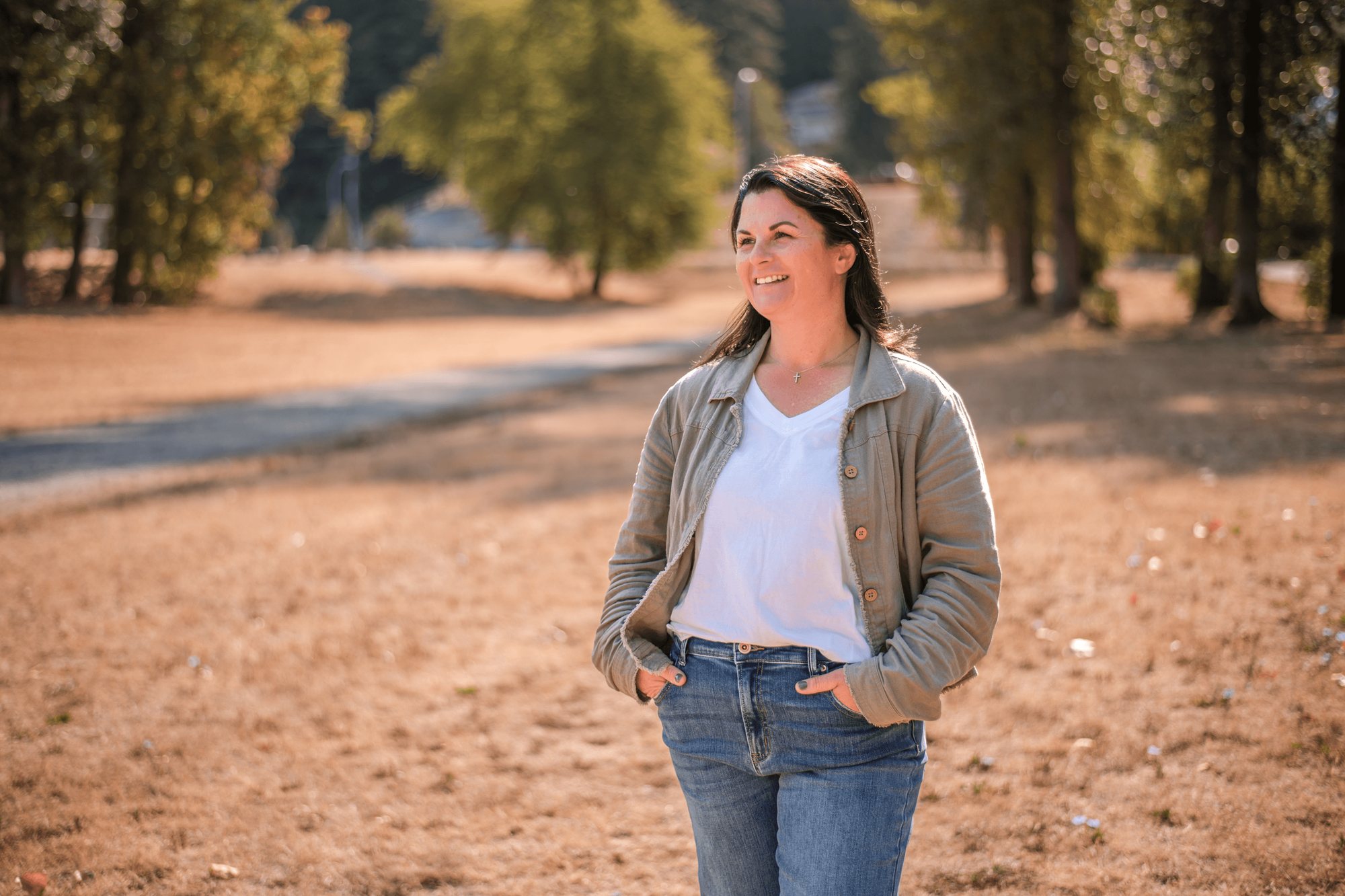 Carey Jones walking across a dry field in a green jacket, white top and jeans, with trees behind her
