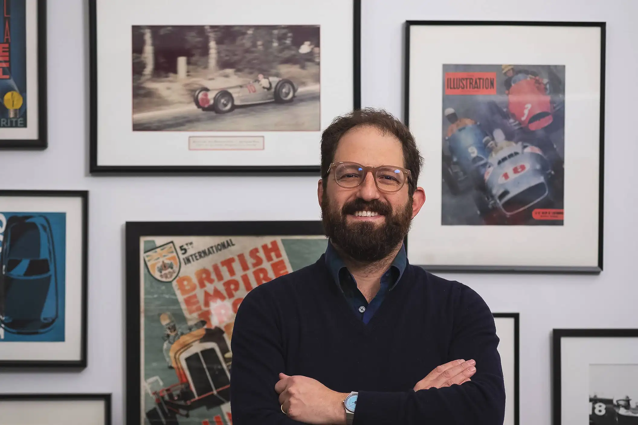 Bradley Price, founder of Autodromo, standing and smiling with arms crossed in front of a wall filled with vintage racing art and posters.