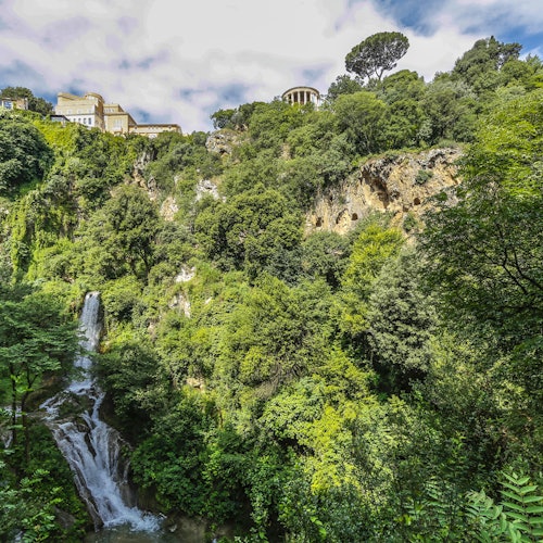 Une cascade dévale des collines verdoyantes et luxuriantes avec des bâtiments perchés au sommet sous un ciel partiellement nuageux.
