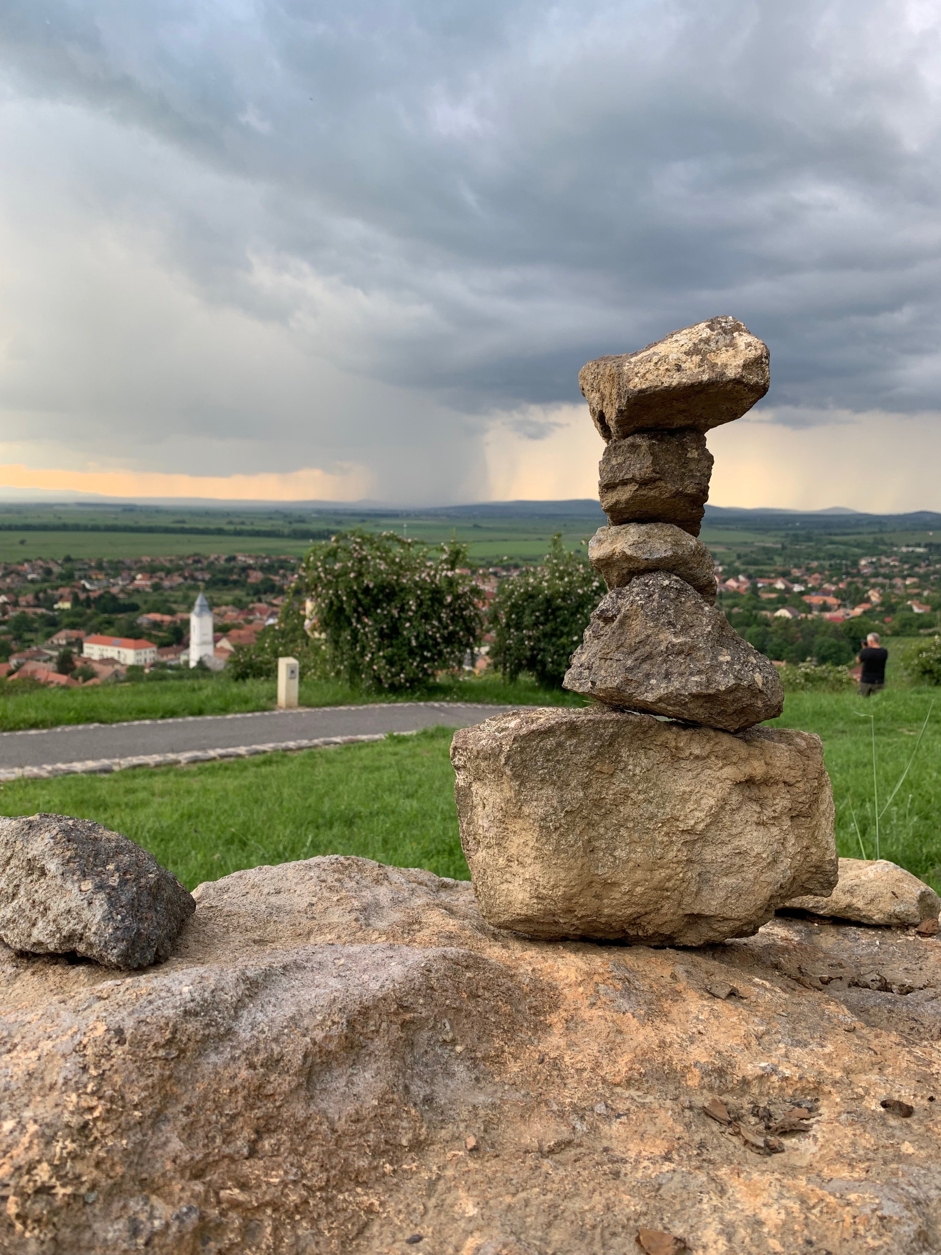 Szabálytalan kövekből álló, gondosan kiegyensúlyozott kőrakás áll egy buja zöld táj előtt, a háttérben egy kisváros távoli látképével felhős ég alatt.