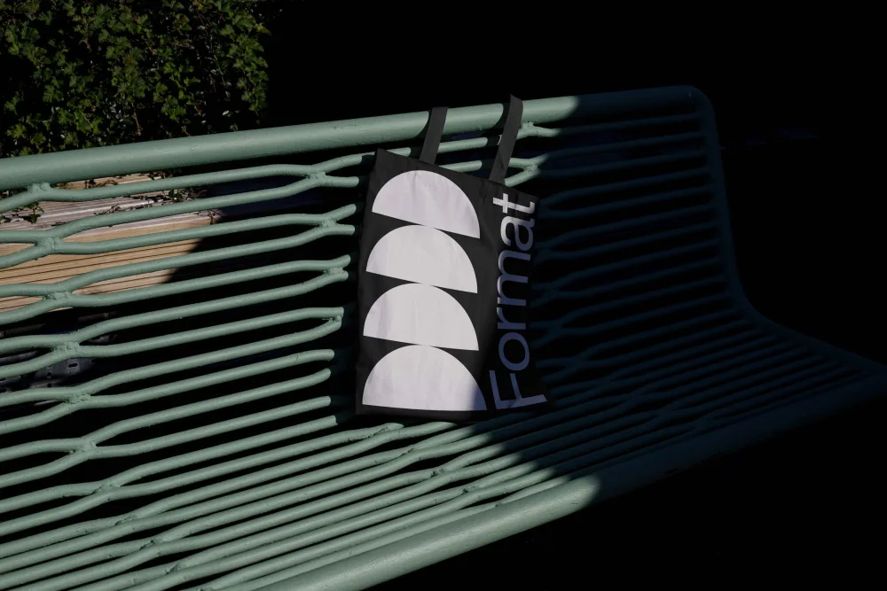 A white tote bag with a bold black radial design rests on a vibrant green metal bench. The scene is illuminated by natural sunlight, creating sharp contrasts and deep shadows, with greenery in the background.