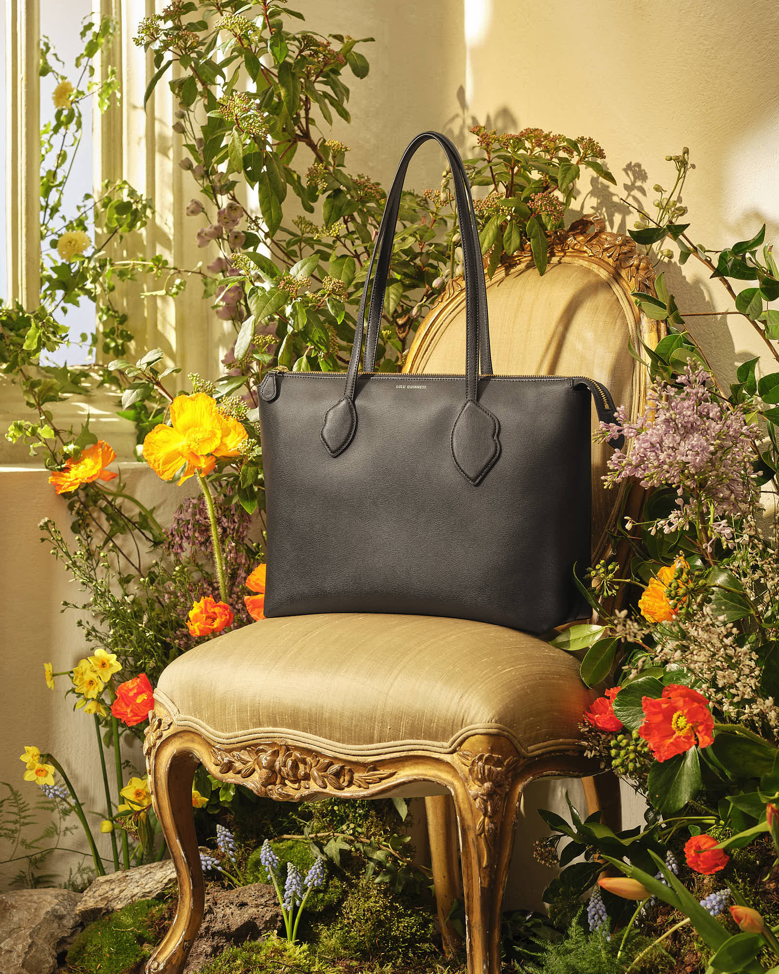 Elegant black leather handbag on vintage chair surrounded by vibrant flowers and greenery.