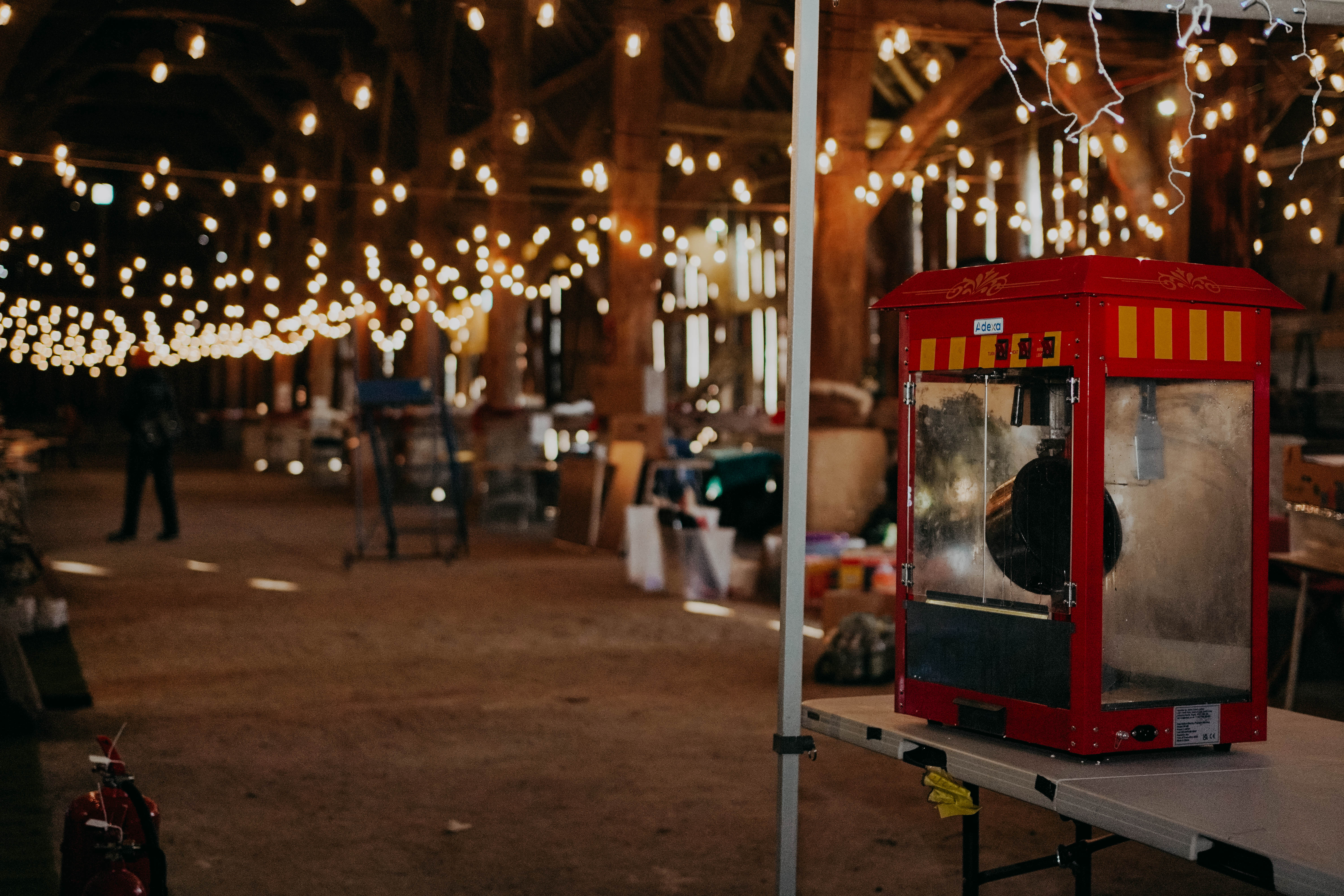 A red popcorn machine sits on a table inside a rustic barn, which is warmly illuminated by numerous strings of glowing lights hanging from the high wooden rafters.