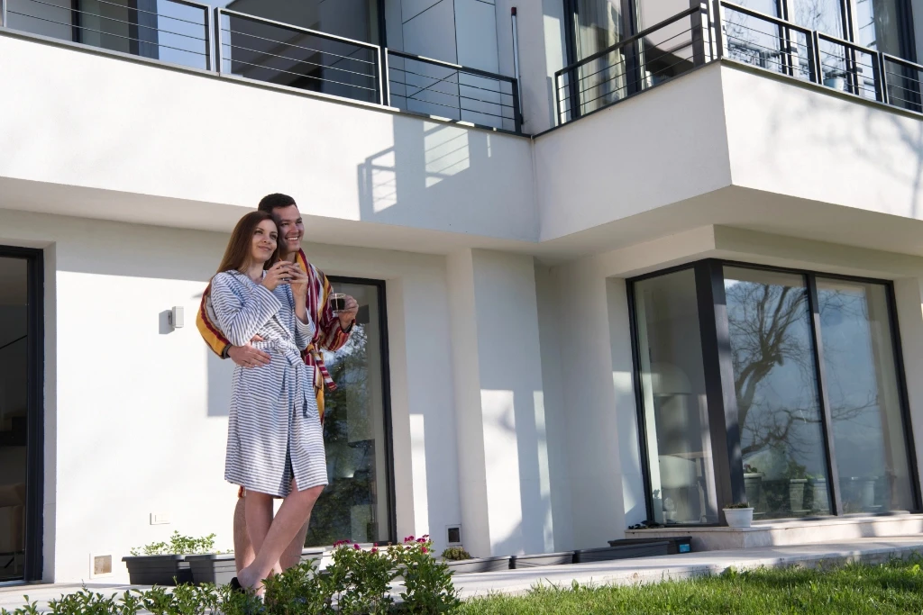A couple standing outside their home, wearing robes and drinking coffee.