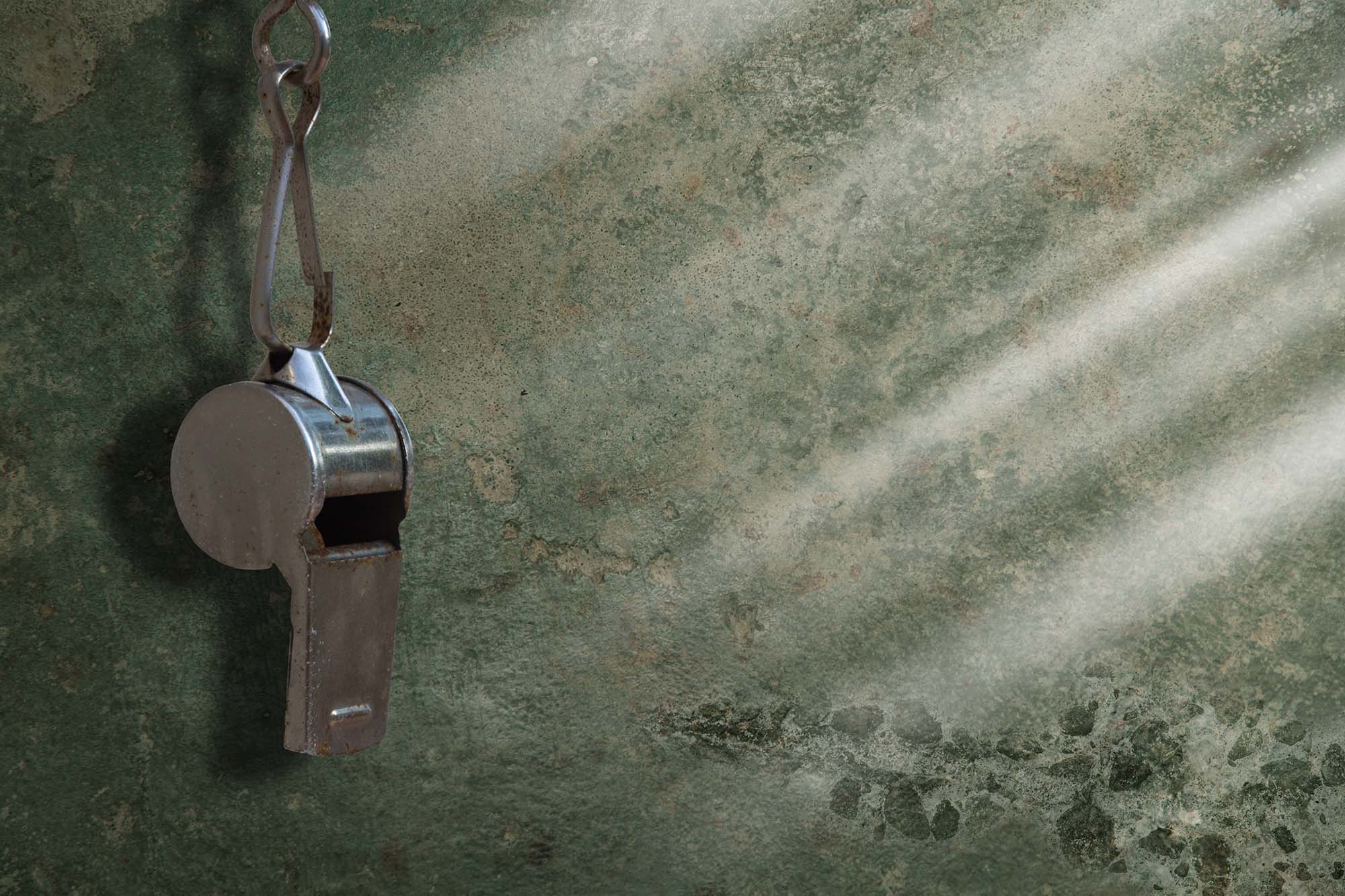 A vintage metal referee whistle hanging on a carabiner clip against a worn green surface, with light rays emanating from its opening.