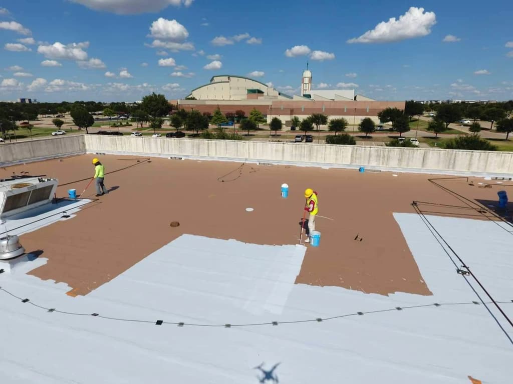 Workers apply white coating to a large flat roof with brown patches.
