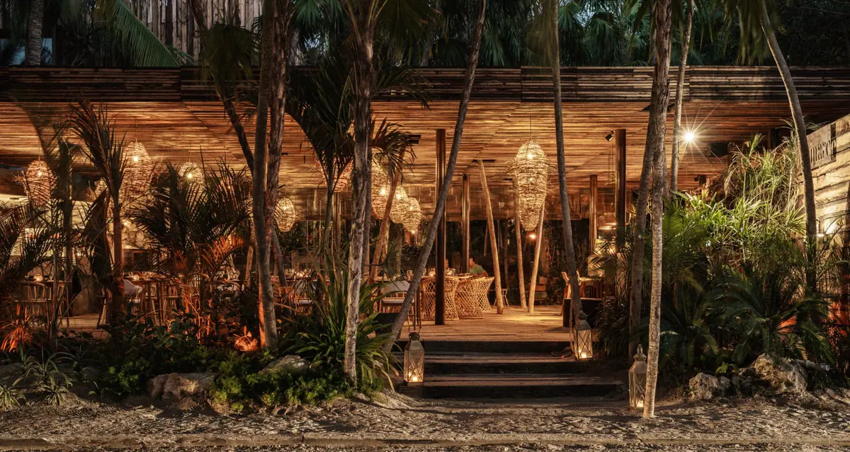 Restaurante Ocumare en Be Tulum, que sirve cocina mexicana, rodeado de exuberante vegetación y ambiente selvático, bien iluminado al anochecer con lámparas colgantes.