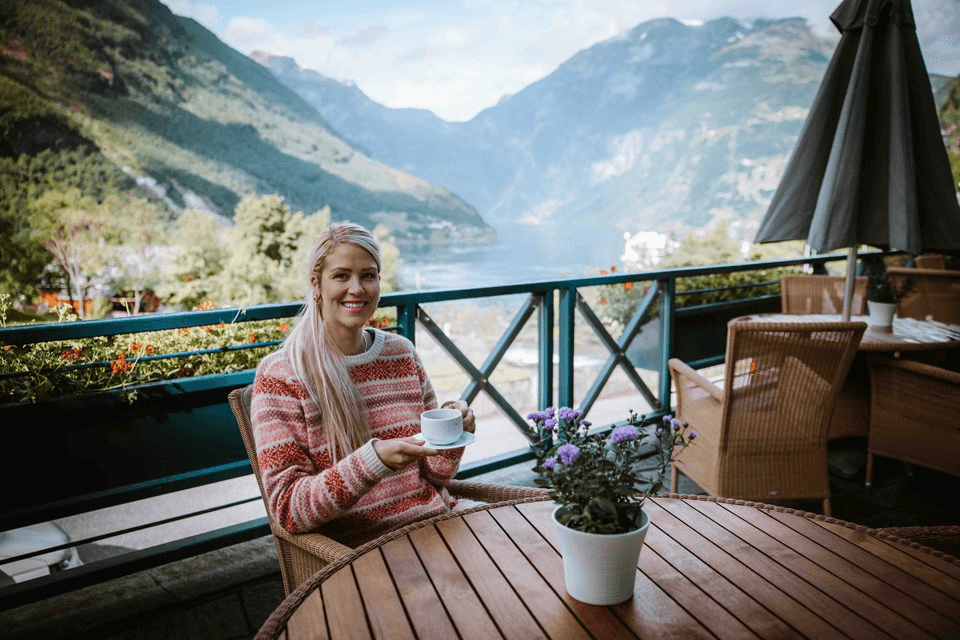 A woman sits at a table with a flower pot, enjoying a scenic mountain view in a cozy outdoor setting.