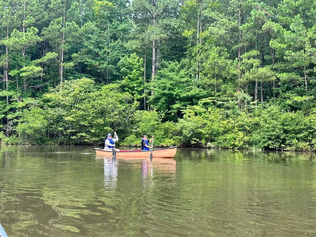 Two people canoeing on a calm lake, surrounded by lush green trees.