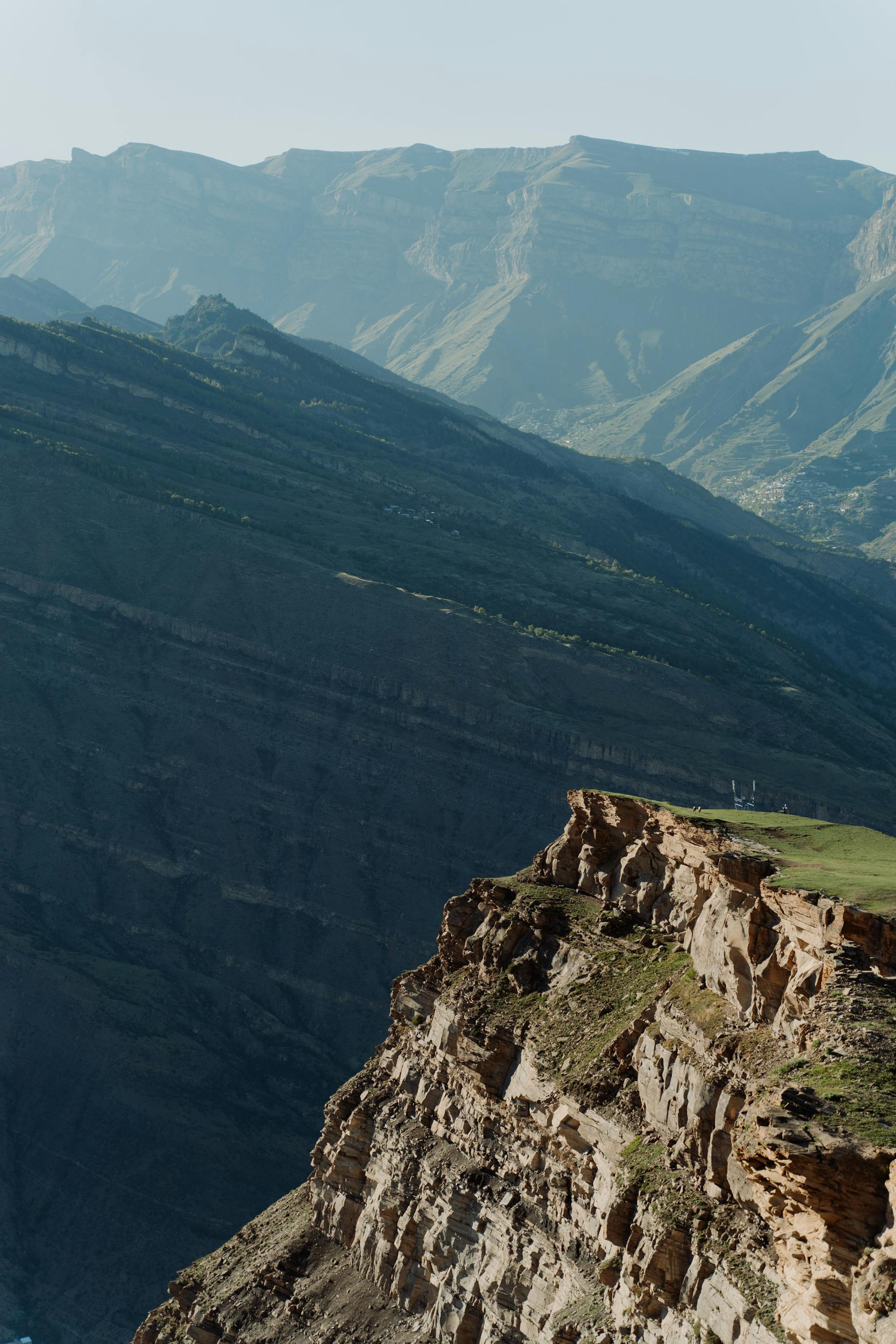 A sheer, rocky cliff edge with a small patch of green grass overlooks a deep valley and massive, sunlit mountain ridges in the distance.