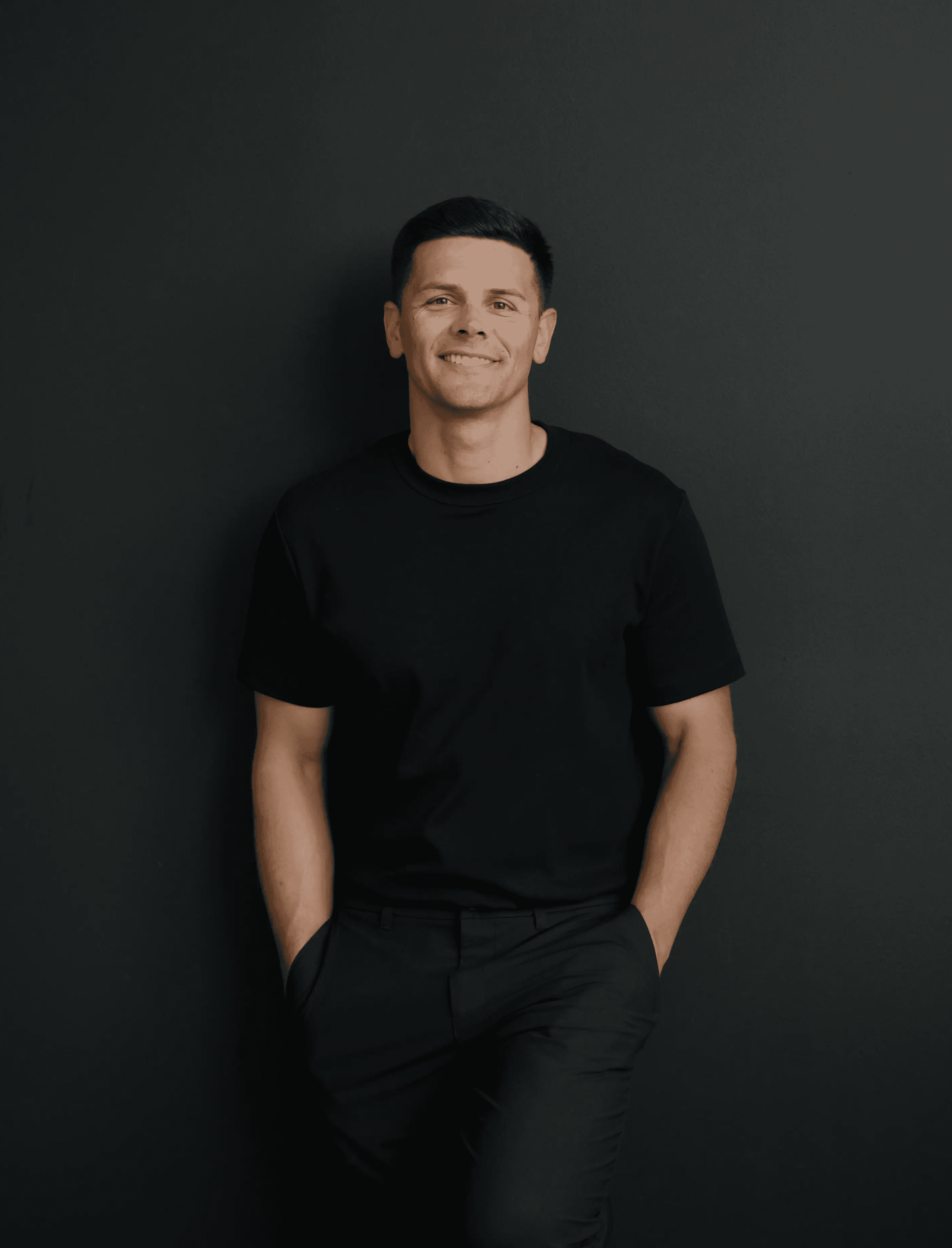 A wedding videographer wearing a black shirt and pants stands casually with hands in pockets against a dark background.