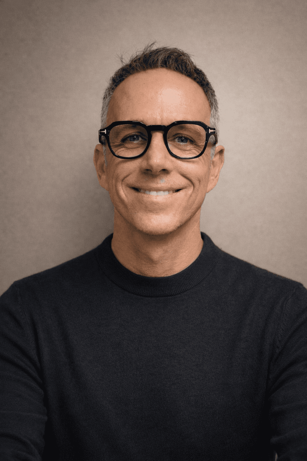 A smiling man with short hair stands with arms crossed, wearing a dark shirt against a neutral background.