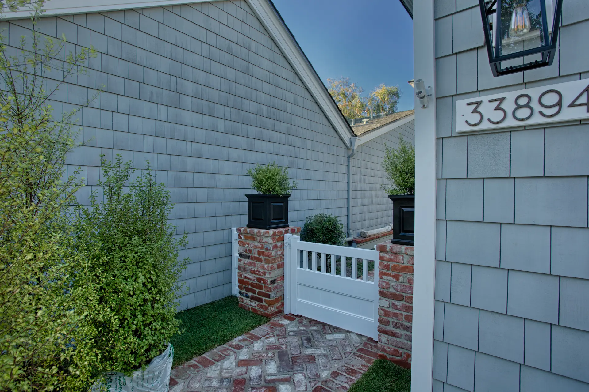 Side entry with brick pathway, white gate, flanked by potted plants and a Cape Cod style home. Photo by Chris Darnall.