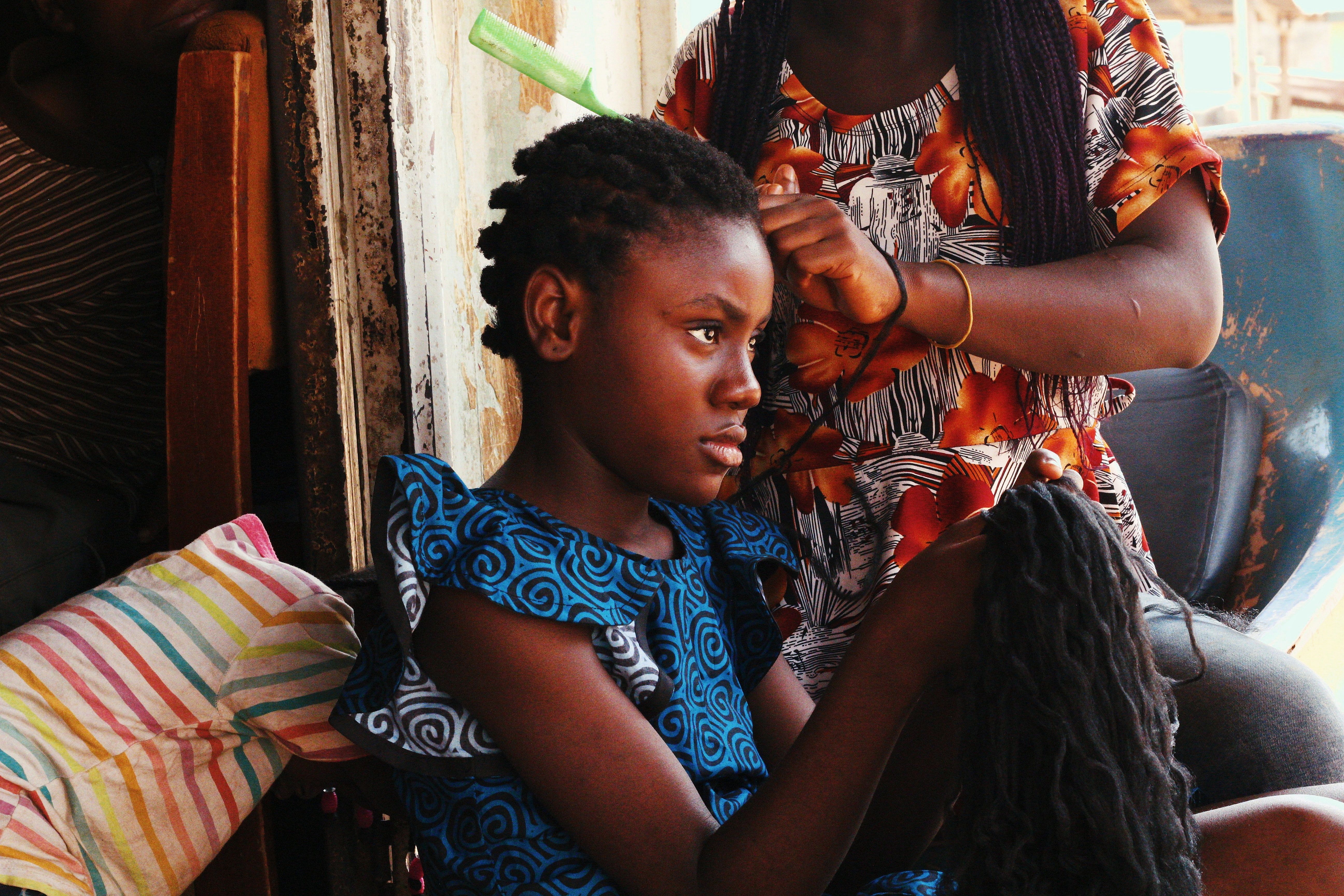 A young girl in Abuja, Nigeria, gets her hair braided while seated indoors. Muhammad-Taha Ibrahim.