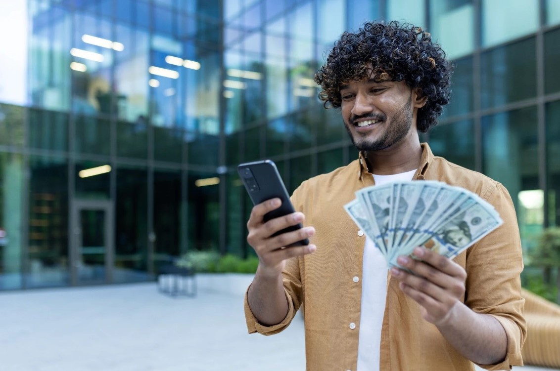 Man holding a phone in one hand and money in the other while smiling. 