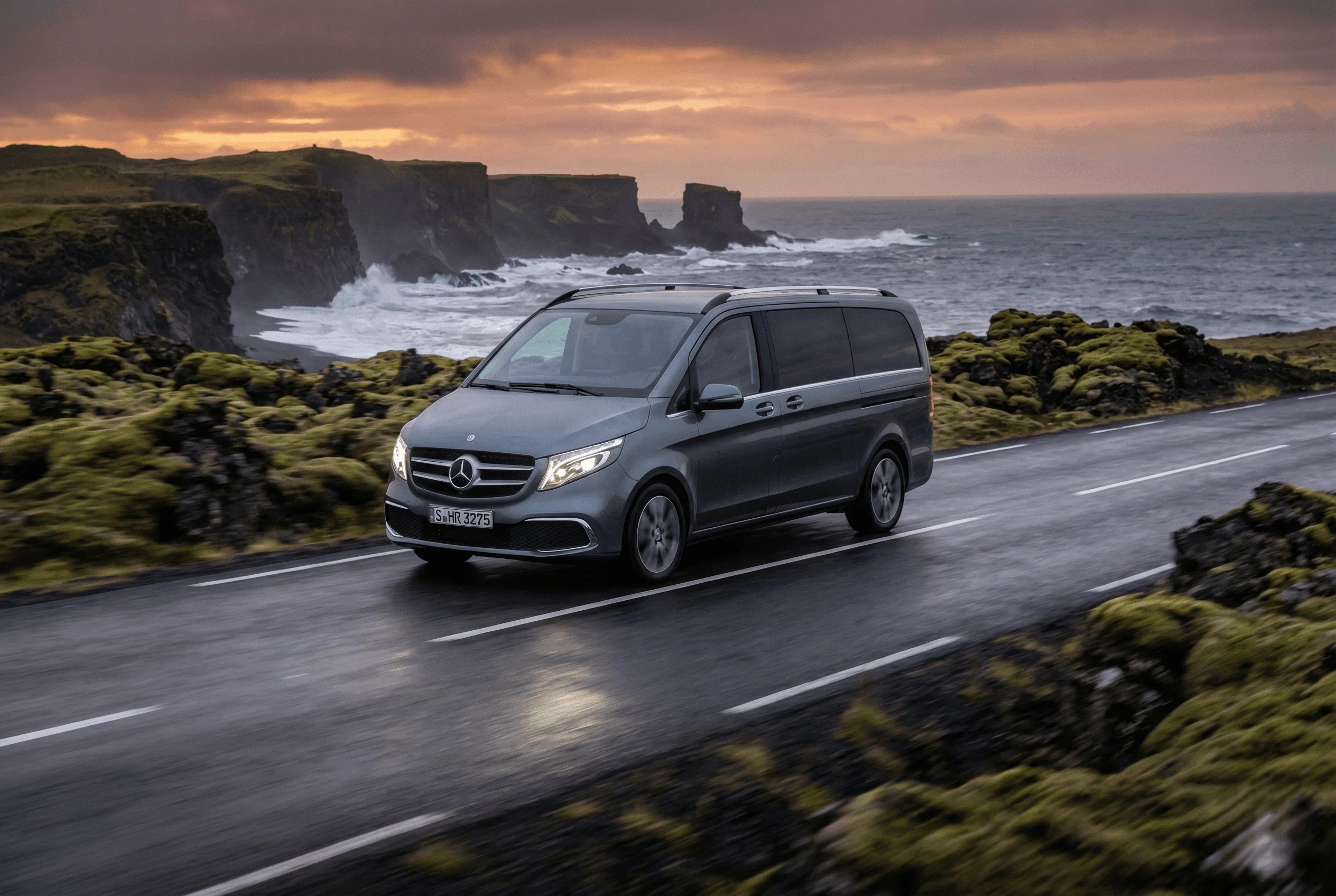 A silver Mercedes van driving along a coastal road with cliffs and the ocean in the background at sunset.