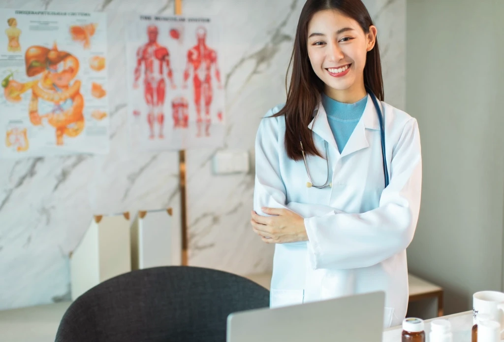 Asian Female doctor standing smiling at camera, with medical diagrams on the wall in the background.