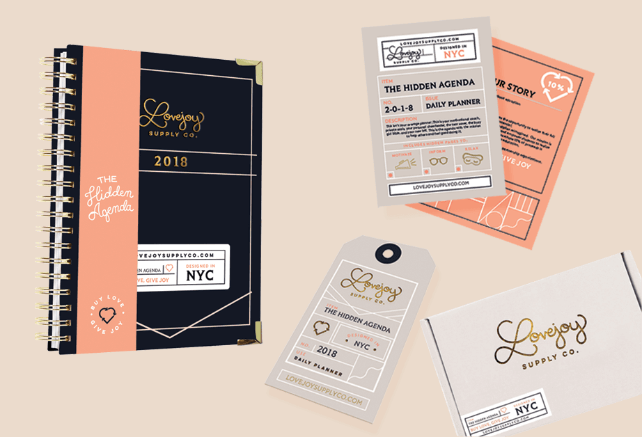 The image shows a collection of stationery and planners from Lovejoy Supply Co., including a dark navy spiral-bound planner from 2018 with a peach-colored band displaying “THE HIDDEN AGENDA,” a daily planner with the same branding, a tag displaying “THE HIDDEN AGENDA NO. 2018 NYC,” and a white envelope with the Lovejoy Supply Co. logo. A second planner, “THE HIDDEN AGENDA DAY PLANNER,” is also visible. All items feature the company’s gold logo and branding. The background is a light beige color.