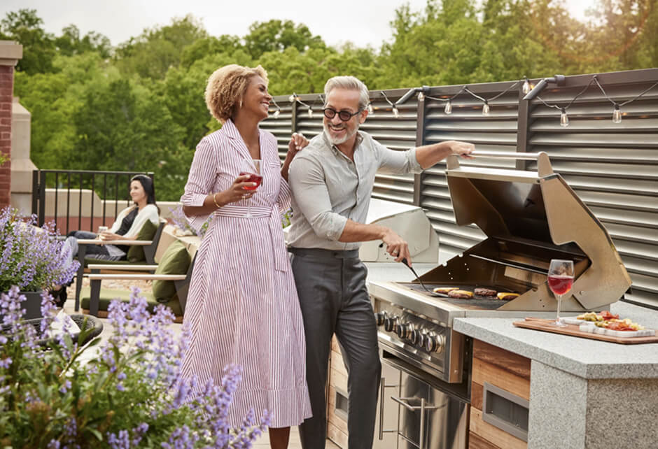 A man grills food on an outdoor barbecue while a woman stands beside him holding a drink; other people sit in the background.