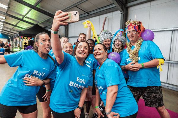 Outdoor team-building event with participants wearing matching blue shirts, taking part in inflatable obstacle courses and games, smiling and cheering, with the words 'ENTER A TEAM' in large bold white and green text across the image.