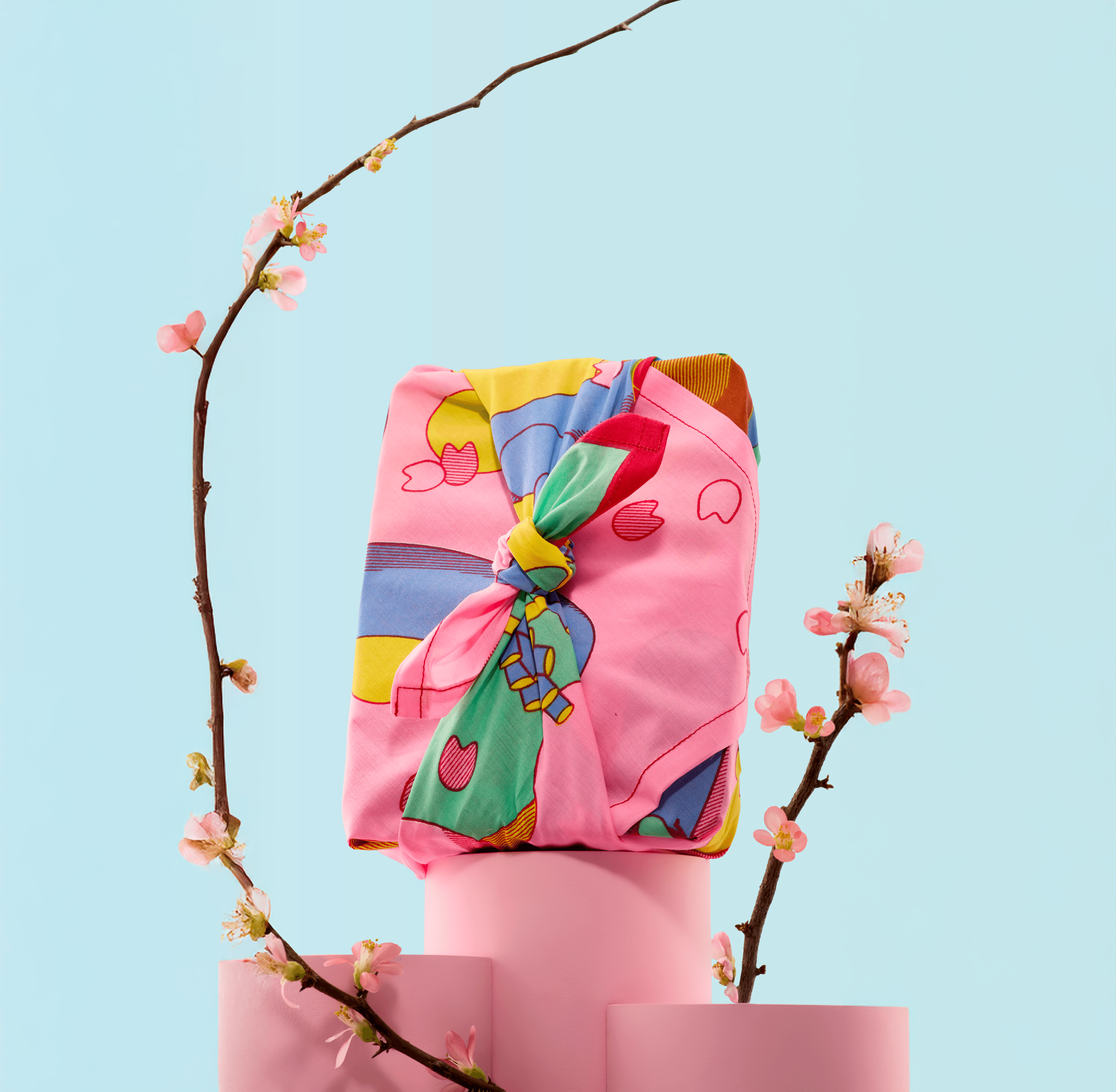 Pink gift box wrapped with a colorful Knot Wrap, tied at the top and displayed on stacked pink blocks with cherry blossom branches.