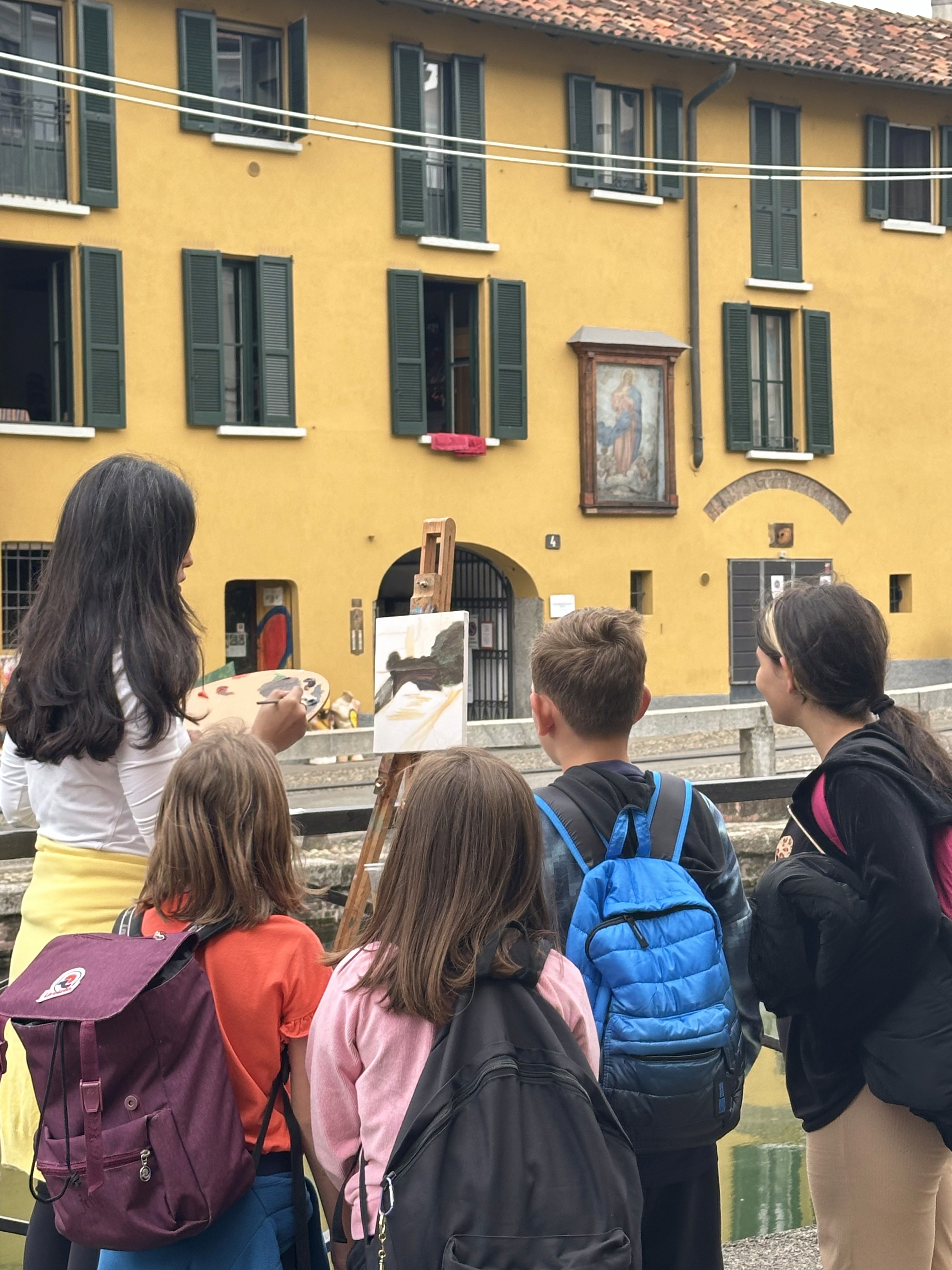 A woman with long dark hair is painting on an easel outdoors, watched by a group of children with backpacks against the backdrop of a yellow building with green shutters and canal water reflecting below.