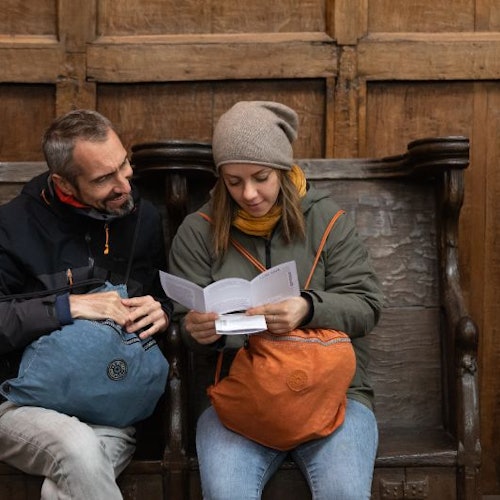 Dua orang duduk di bangku kayu. Yang satu membaca buku panduan sementara yang lain melihat. Keduanya membawa ransel.