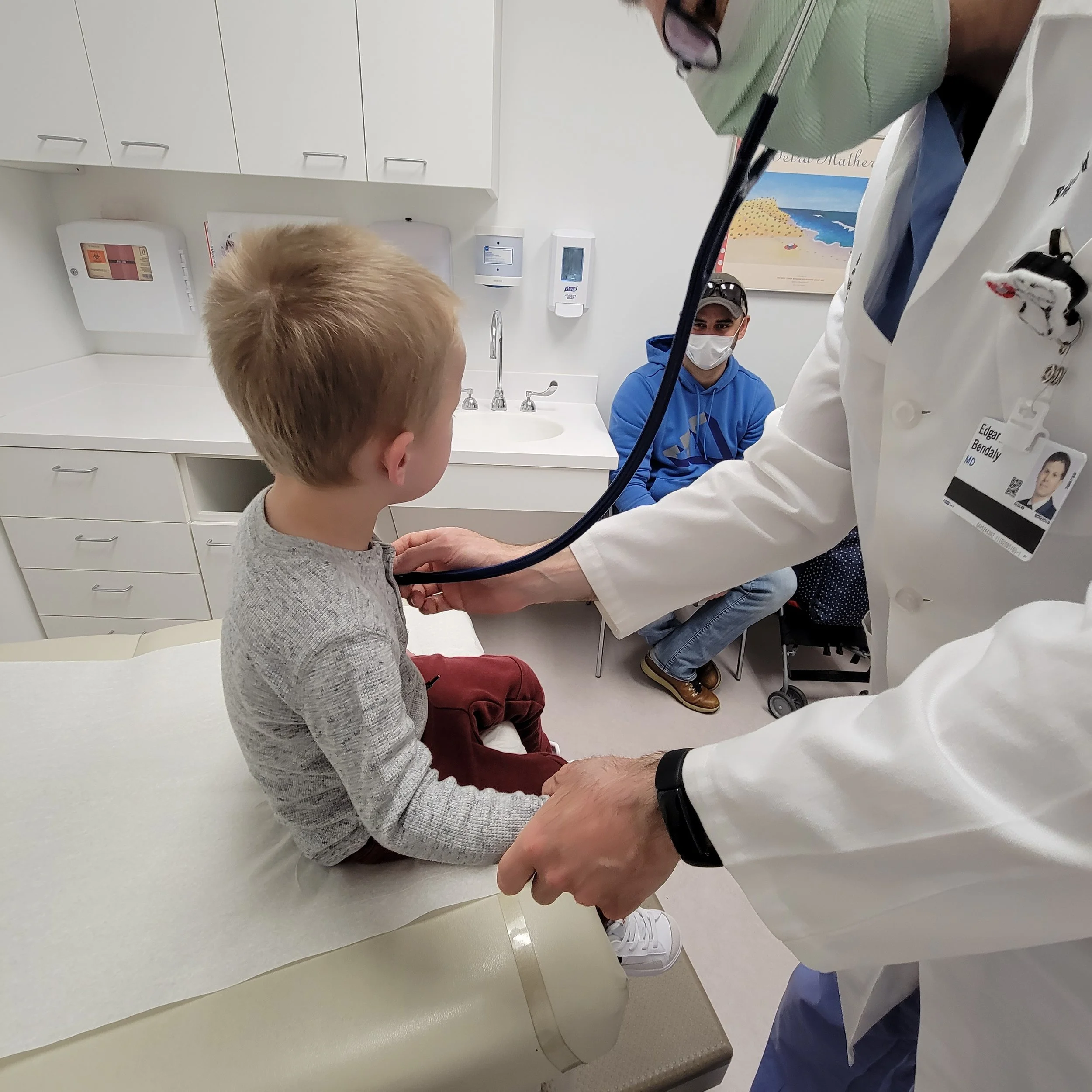 pediatric patient sitting on exam table in doctor's office while doctor listens to heart and lungs with stethoscope and parents sit by the side