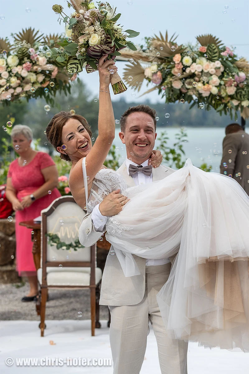 Lächelnder Bräutigam trägt Braut, Schloss Mattsee, mit Brautstrauß. Bubbles.