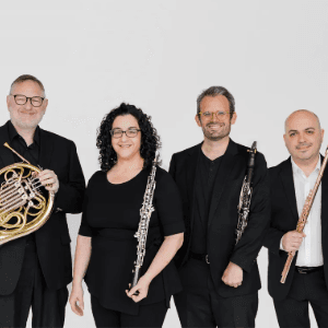 Headshot of four musicians in black. A man holding a french horn, a woman holding an oboe, a man holding a clarinet, and a man holding a flute.