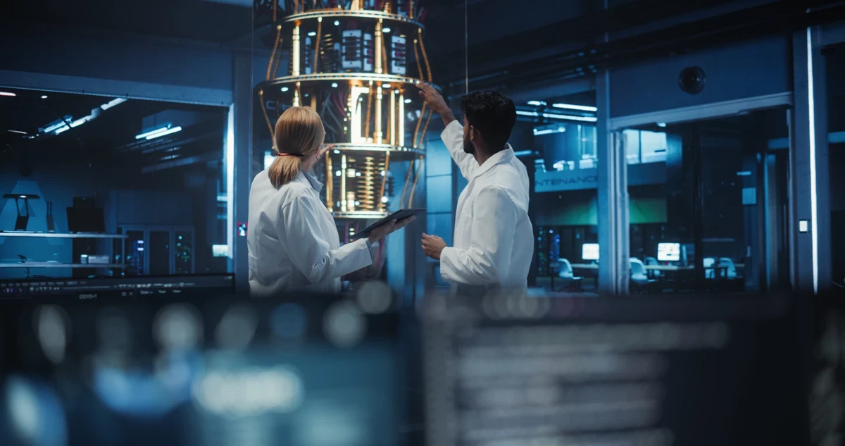 Two people in lab coats examining a large cylindrical quantum computing device in a high-tech laboratory, with computer screens visible in the foreground.