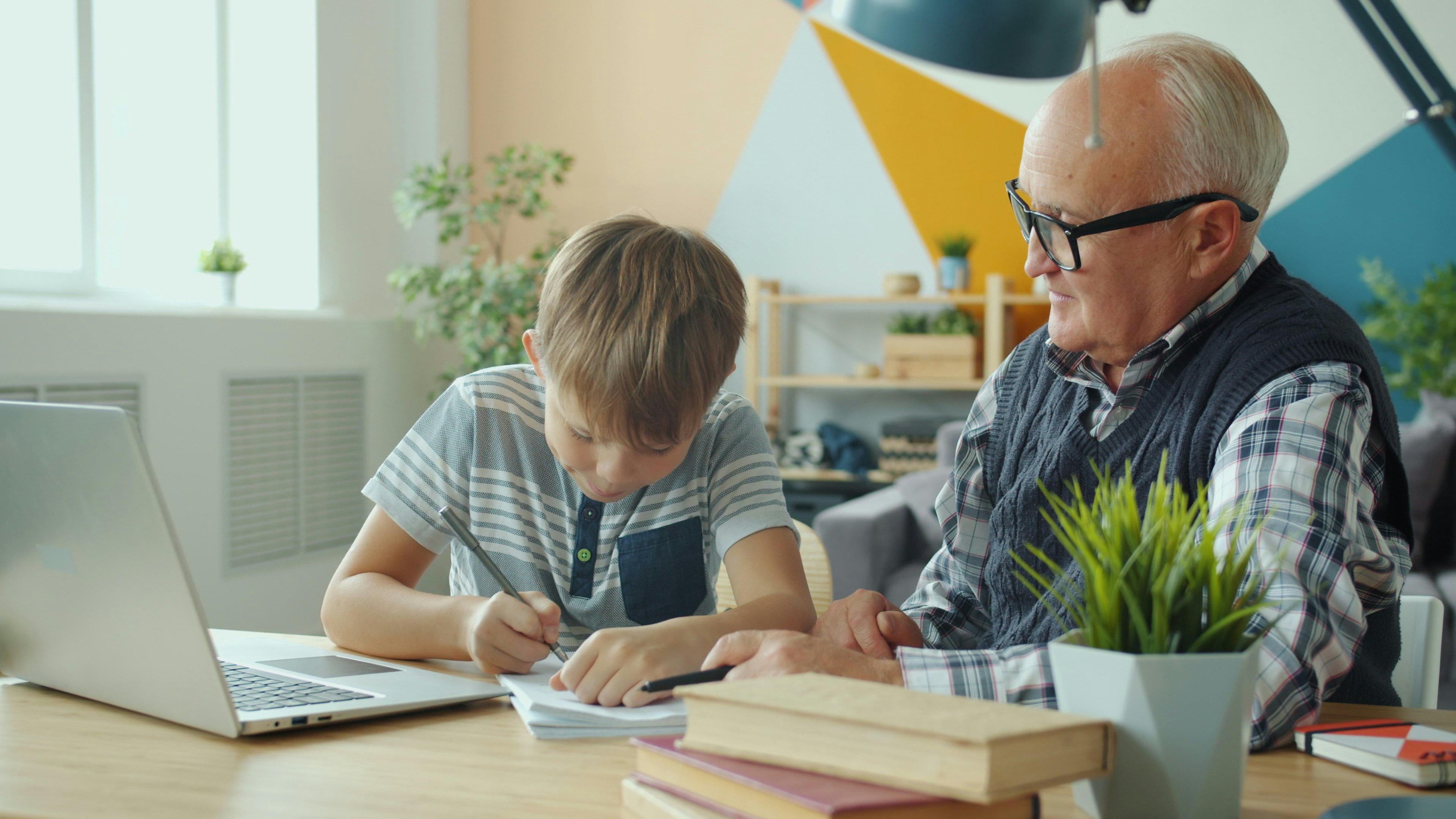 An older man sits beside a boy at a table while the boy writes on paperwork next to an open laptop and books.