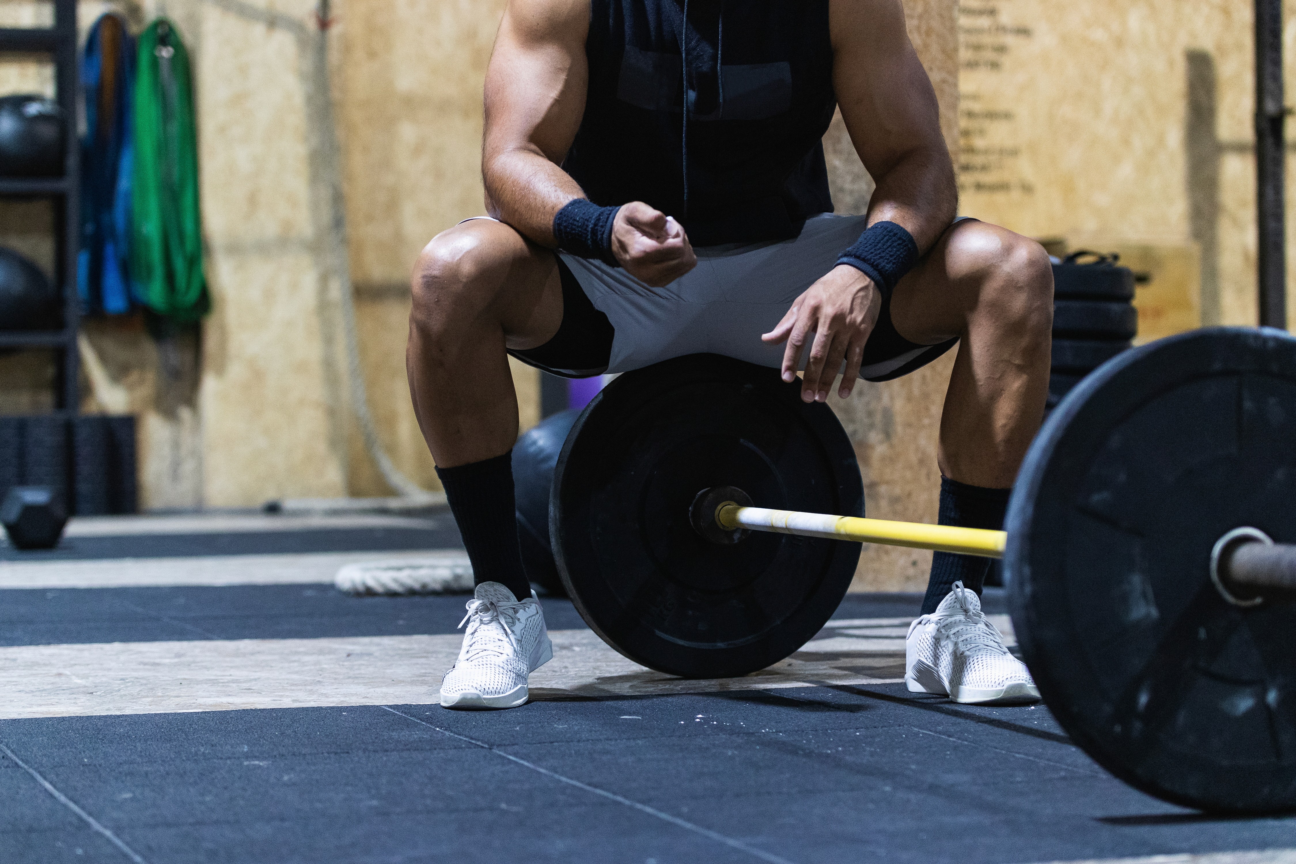 Man resting in a squat position in a gym near a loaded barbell, wearing athletic gear including wristbands and white sneakers. 