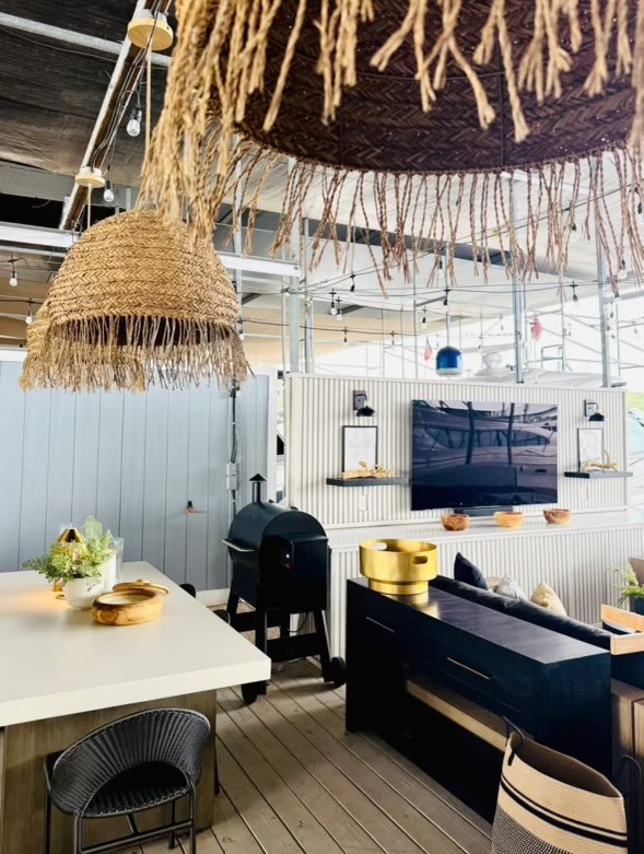 A modern outdoor living space features a cozy seating area, large woven pendant lights, a sleek flat-screen TV on a patterned wall, and a stylish charcoal grill, all set against a backdrop of natural wood flooring.