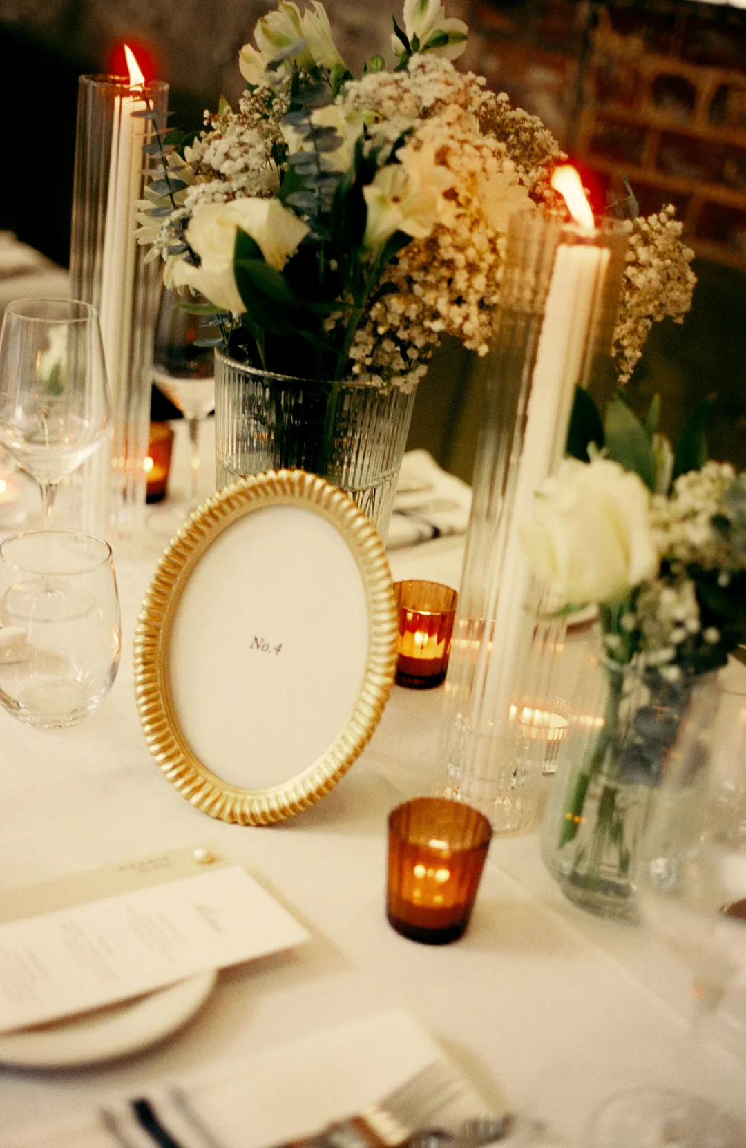Candlelit table with a gold oval frame marked No. 4, tall tapered candles in ribbed glass holders, amber votives and a white floral arrangement with baby’s breath.