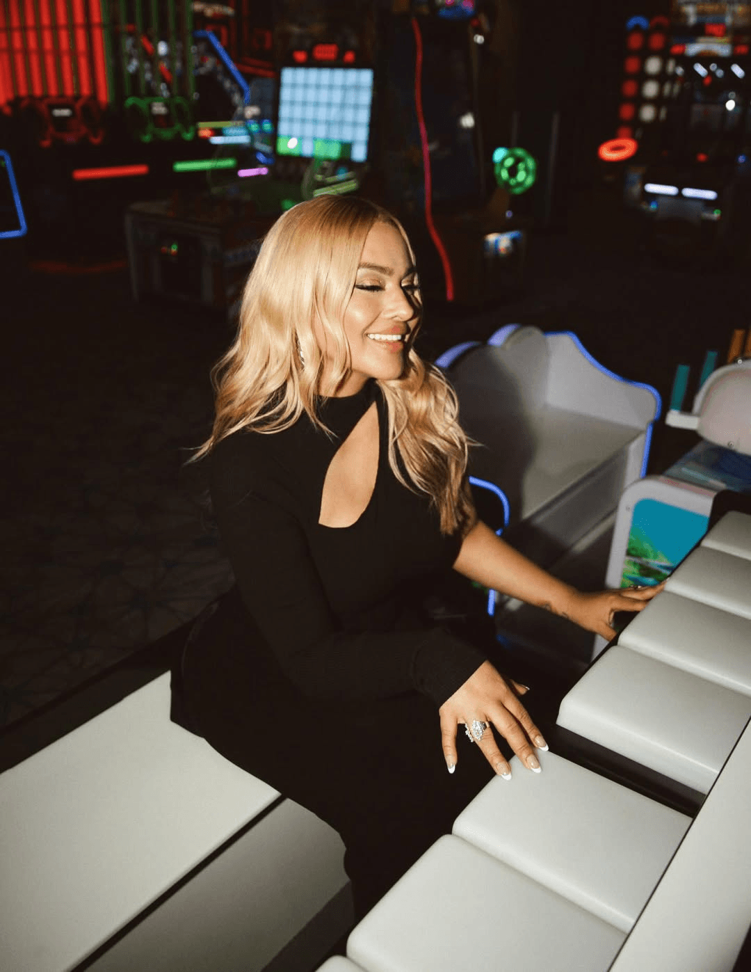A smiling woman with blonde hair stands with her arms crossed, wearing a black shirt against a dark background.