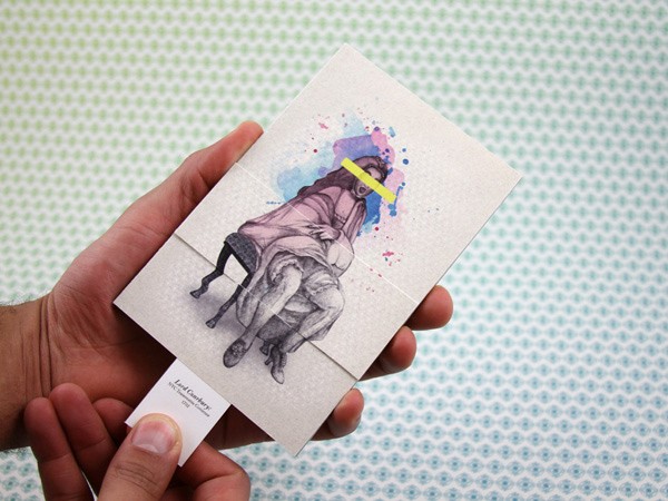 Two hands hold a folded piece of paper with a drawing on it. The drawing depicts two figures, one seated on a chair and the other leaning over them. The figure leaning over has long dark hair and a yellow band across its forehead. The paper is off-white with watercolor splashes in blue, pink, and yellow. A white tag with text is attached to the bottom left corner of the paper. The background is a light teal patterned surface.