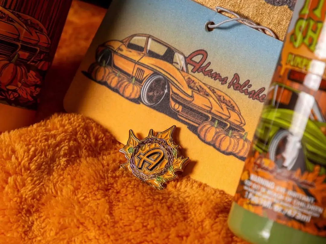 Close-up of Adams Polishes Pumpkin Spice collection artwork featuring illustrated car tag, enamel pin, microfiber towel, and autumn-themed detailing product packaging.