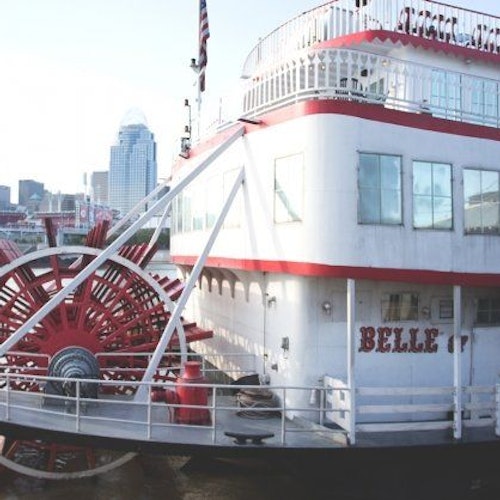 白色蒸汽船“Belle”停靠在码头，船身带有红色饰边，背景是一座城市的天际线。