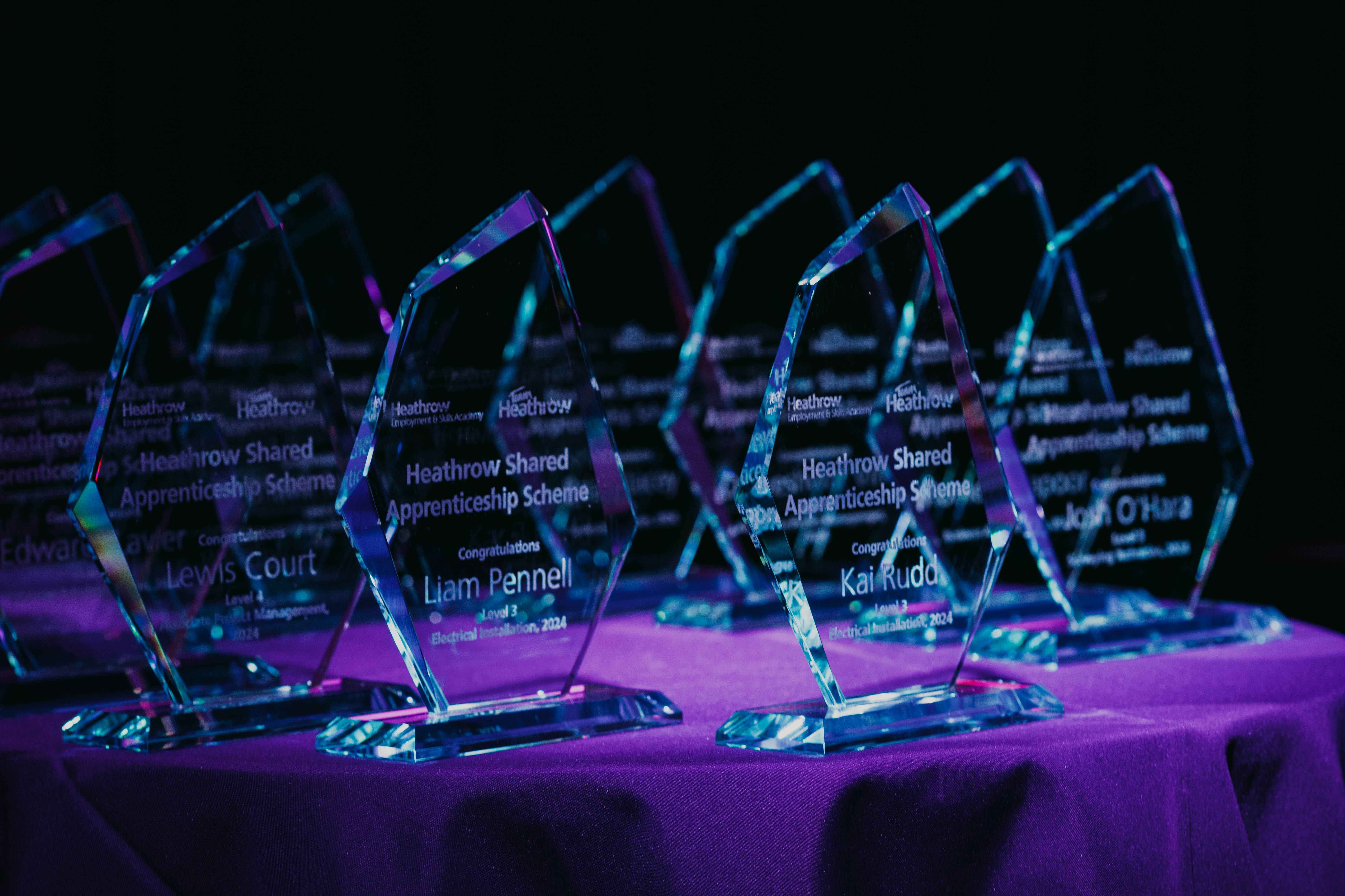 A row of clear, diamond-shaped glass awards are displayed on a purple tablecloth under dramatic lighting. Each award is engraved with the Heathrow logo and text for the "Heathrow Shared Apprenticeship Scheme" and the names of the recipients.