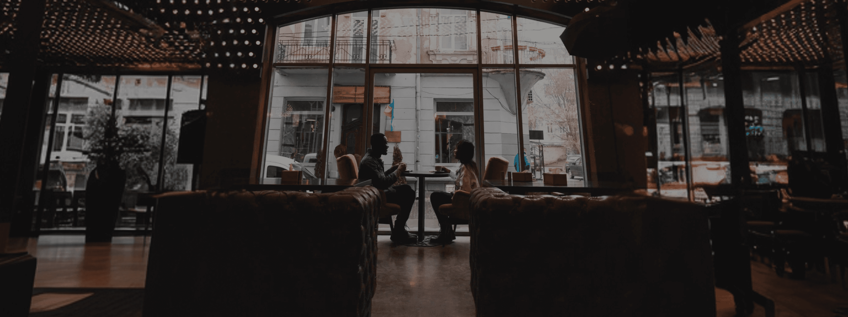 Silhouettes d'un couple discutant autour d'un café dans un intérieur tamisé, avec une vue urbaine à travers une grande baie vitrée.