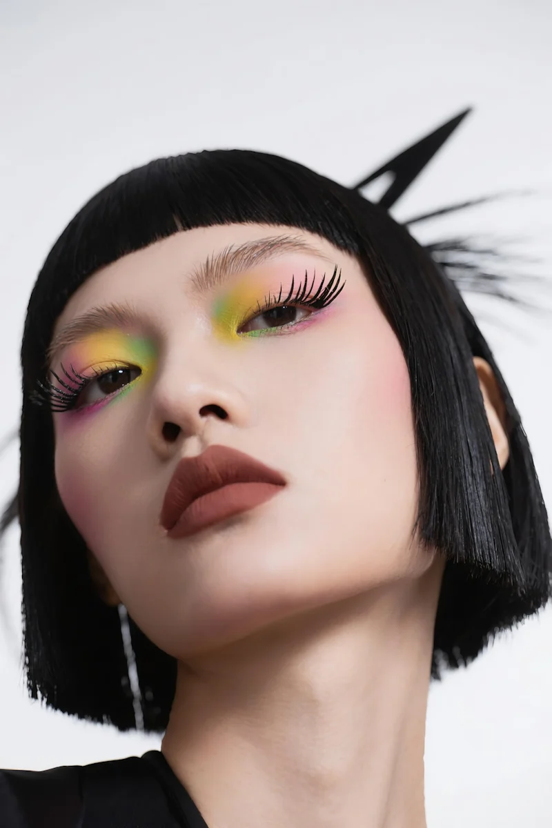 Close-up portrait of a person with bold colorful eyeshadow, long lashes, and a sleek black bob haircut on a white background.