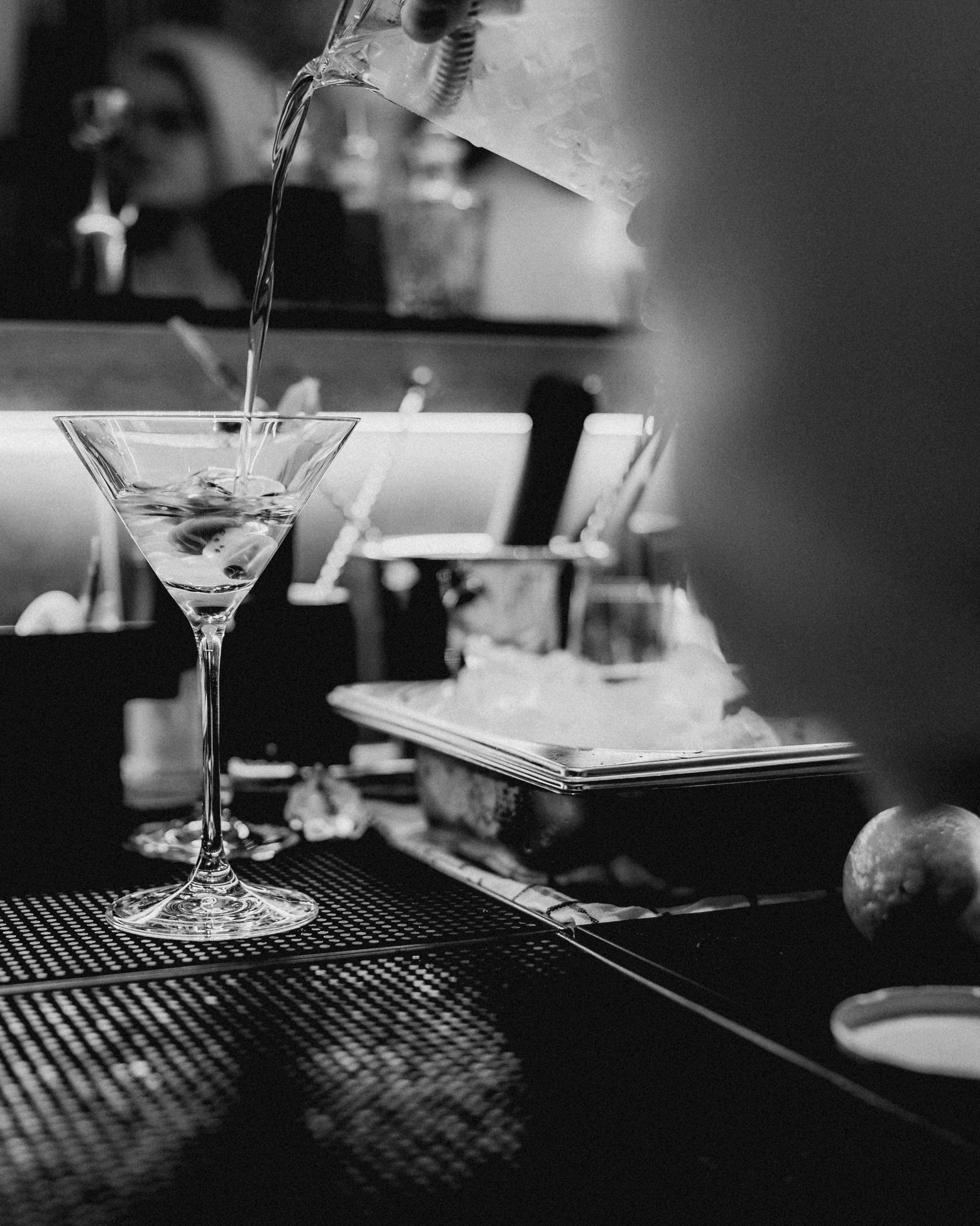 Schwarz-Weiß-Fotografie eines Cocktails, der an einer Bar in ein Martini-Glas gegossen wird