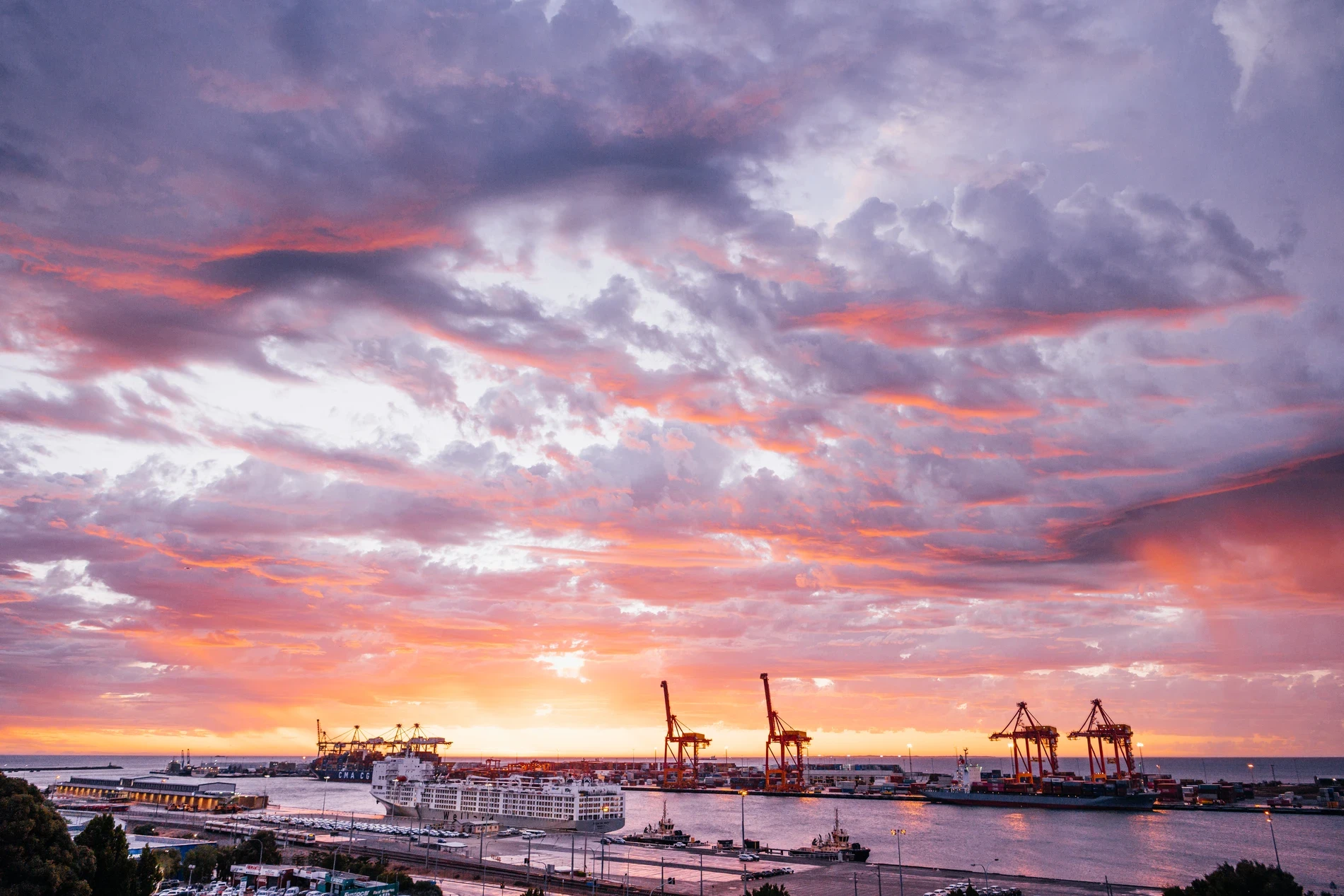 A container port at sunset under a pink and purple sky, cranes silhouetted along the water