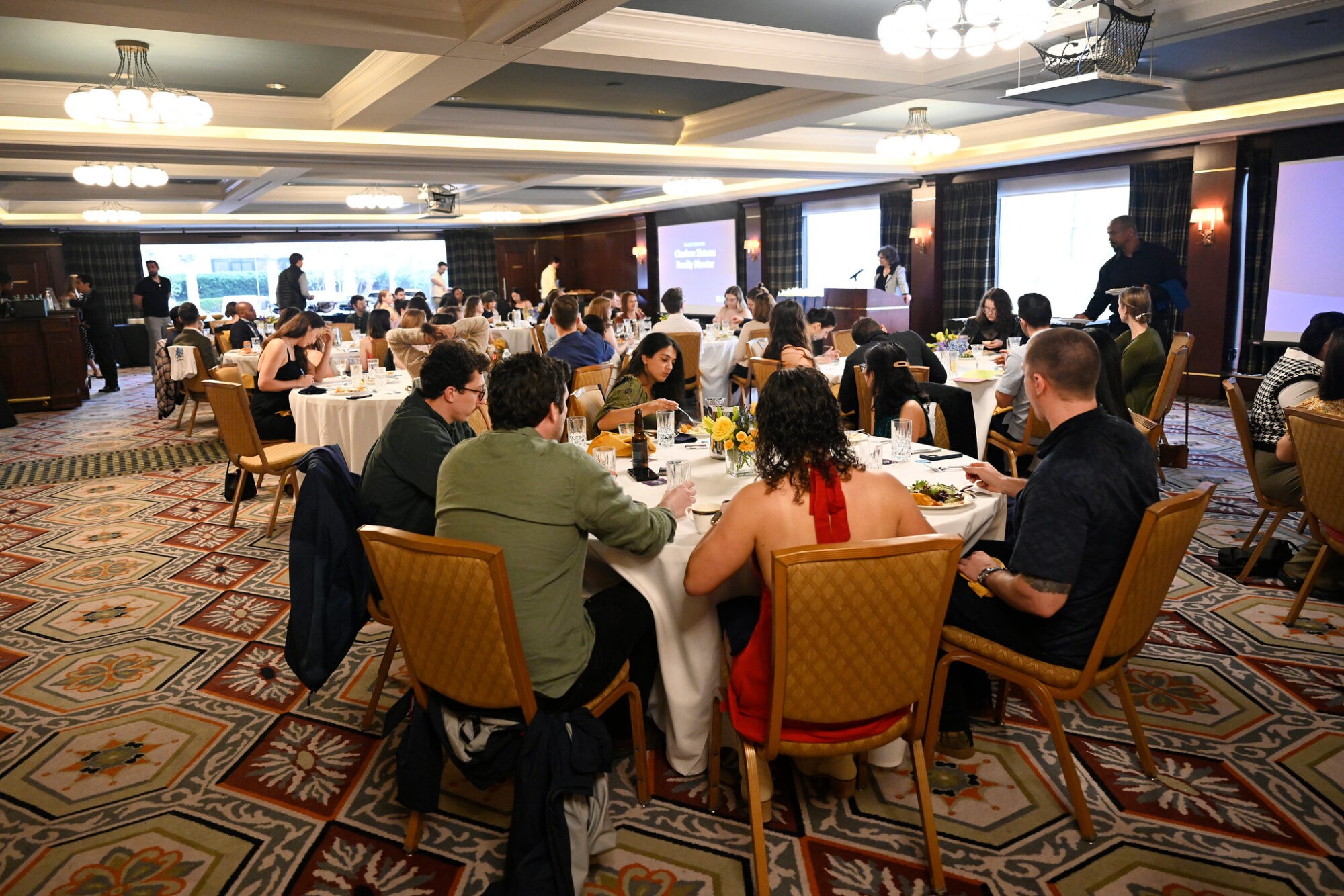 A group of people seated at tables in a conference room, engaged in discussion with presentations in the background.