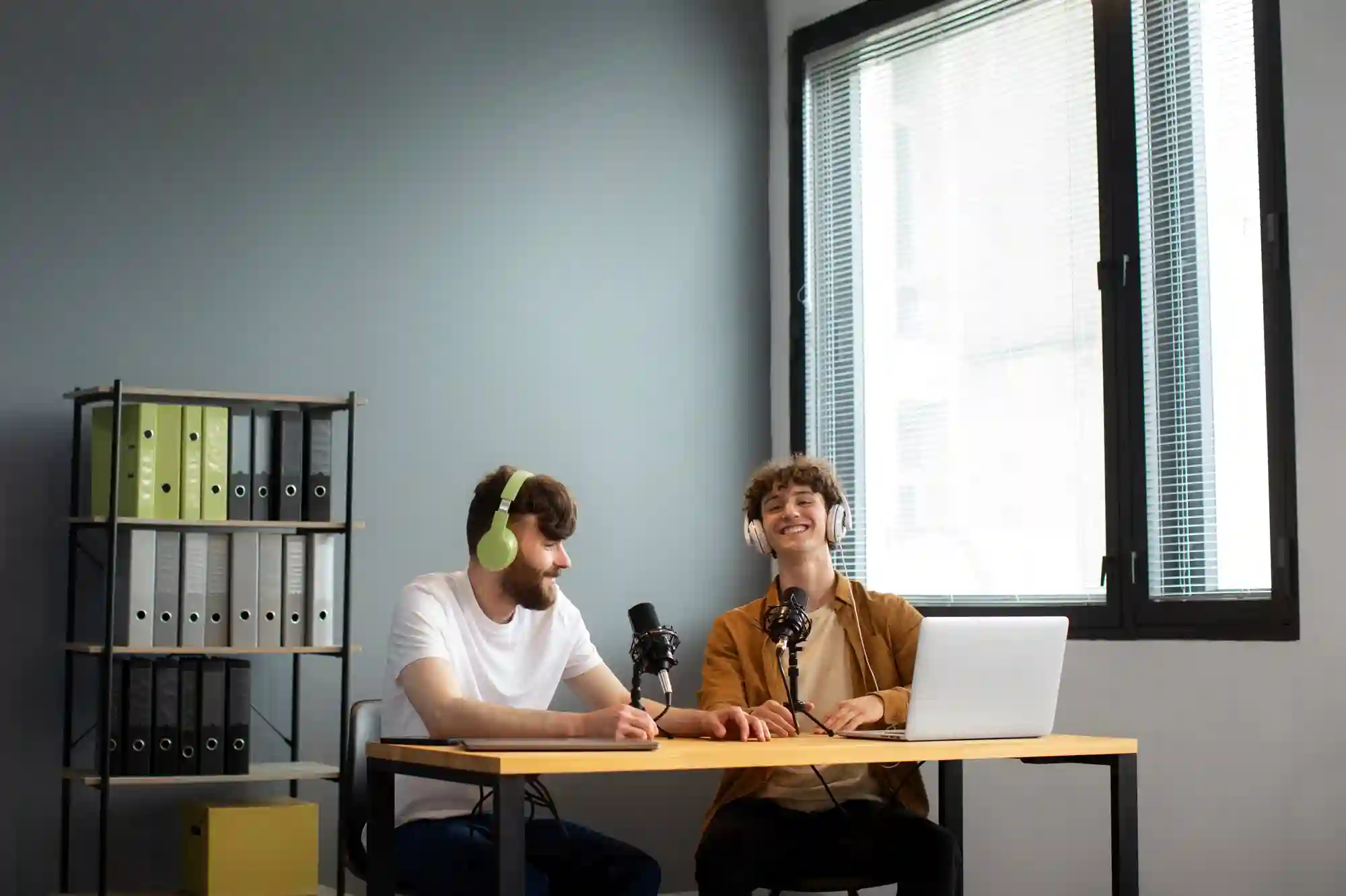 Two men with headphones recording a podcast in a small studio with a microphone and laptop